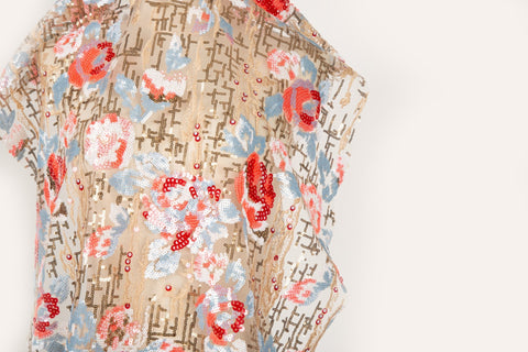 A sequin embroidery fabric on nude mesh with red-blue florals over bronze geometric patterns.

