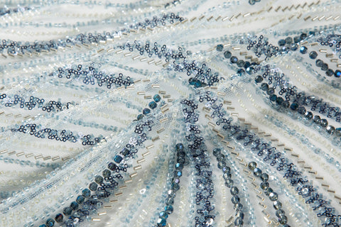 A blue and silver hand-beaded fabric, with an abstract, flowing pattern resembling water currents or mineral strata.
