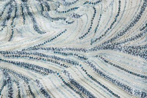 A blue and silver hand-beaded fabric, with an abstract, flowing pattern resembling water currents or mineral strata.
