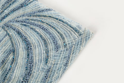 A blue and silver hand-beaded fabric, with an abstract, flowing pattern resembling water currents or mineral strata.
