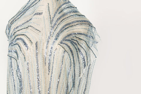A blue and silver hand-beaded fabric, with an abstract, flowing pattern resembling water currents or mineral strata.
