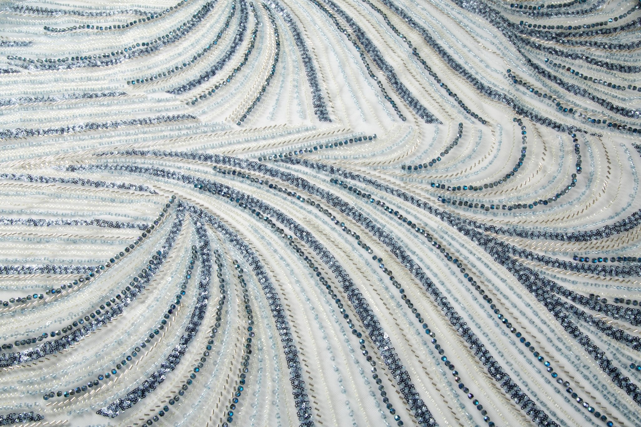 A blue and silver hand-beaded fabric, with an abstract, flowing pattern resembling water currents or mineral strata.

