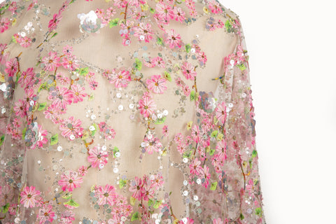 A fine embroidery fabric on white tulle, with a pattern of pink cherry blossom branches and shimmering sequins.

