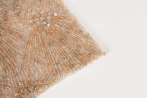 An Art Deco style beaded fabric, with a radiating palm leaf or sunburst pattern in a rose gold and silver palette.
